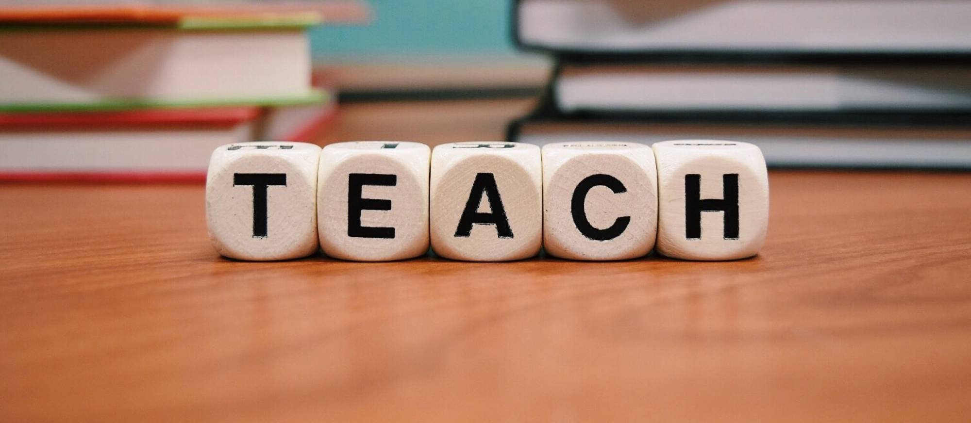 blocks spelling the word teach on a desk with books in the background