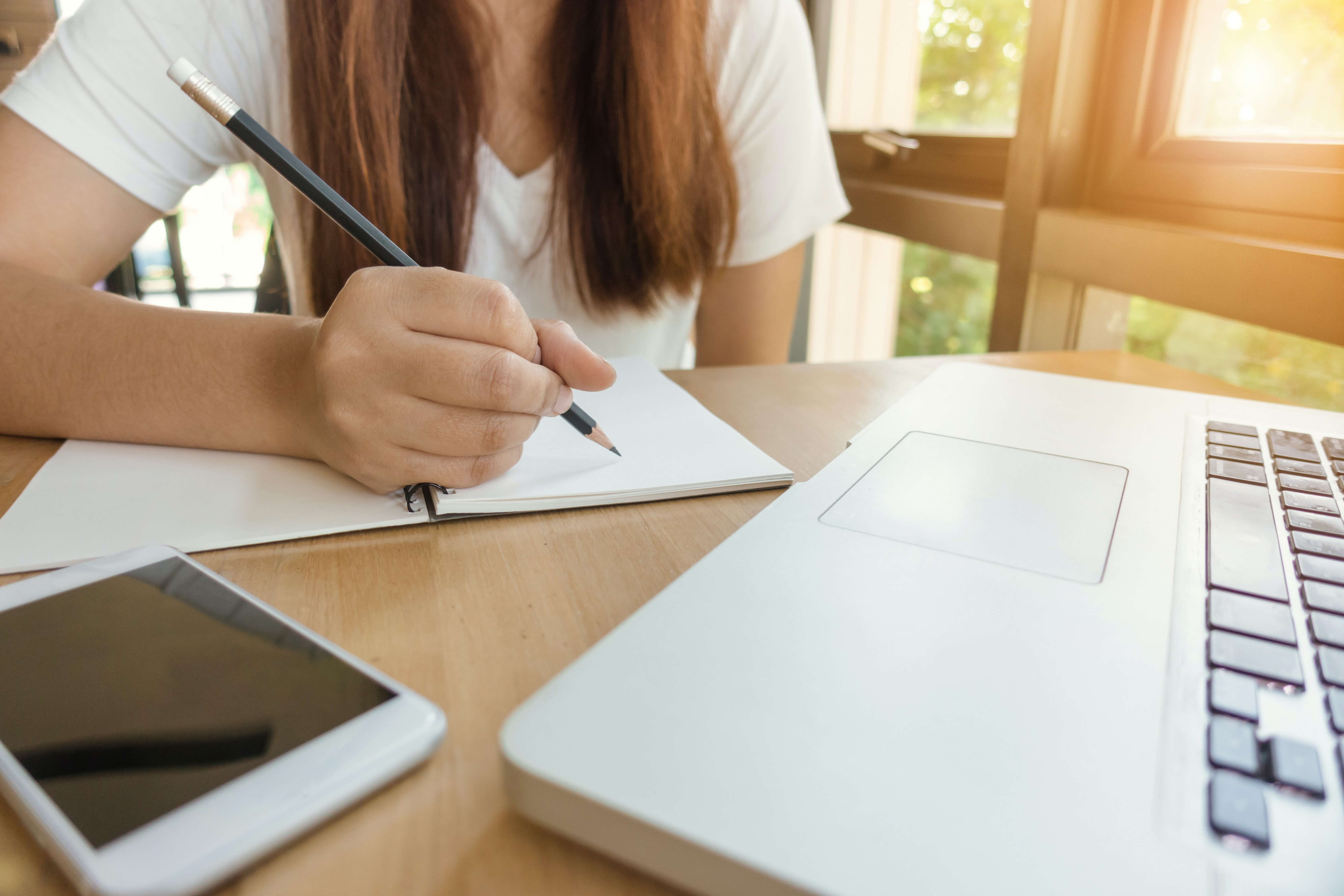 student writing on paper with laptop open